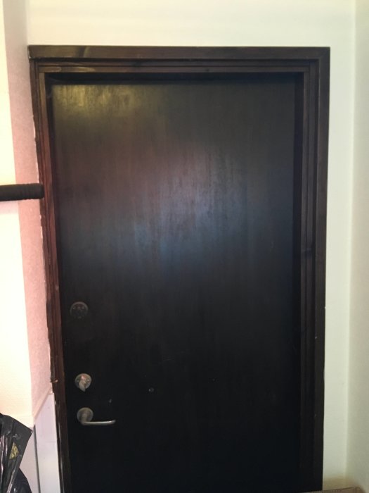En mörkbrun, stängd ytterdörr inomhus ramad av en träkarm, med fråga om isolering och uppsättning av hyllor framför.