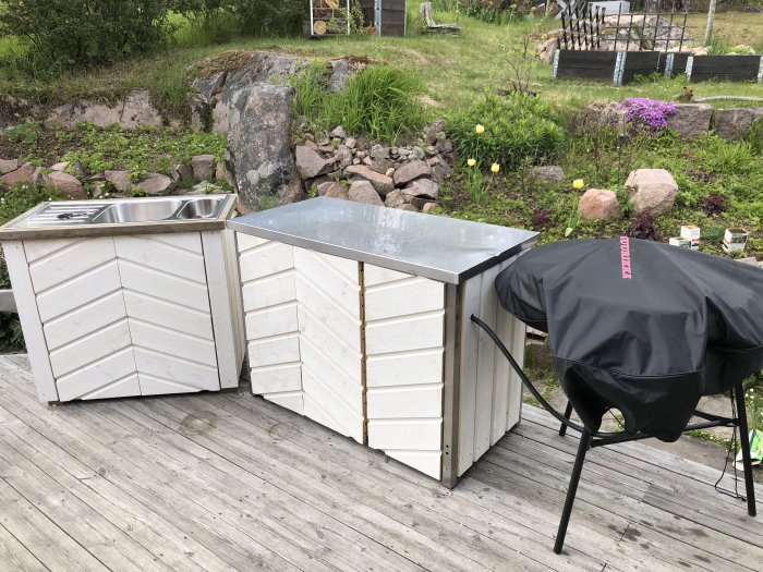 Utekök med rostfritt bord, panelbrädsluckor och en täckt grill på en trädäck med vegetation i bakgrunden.