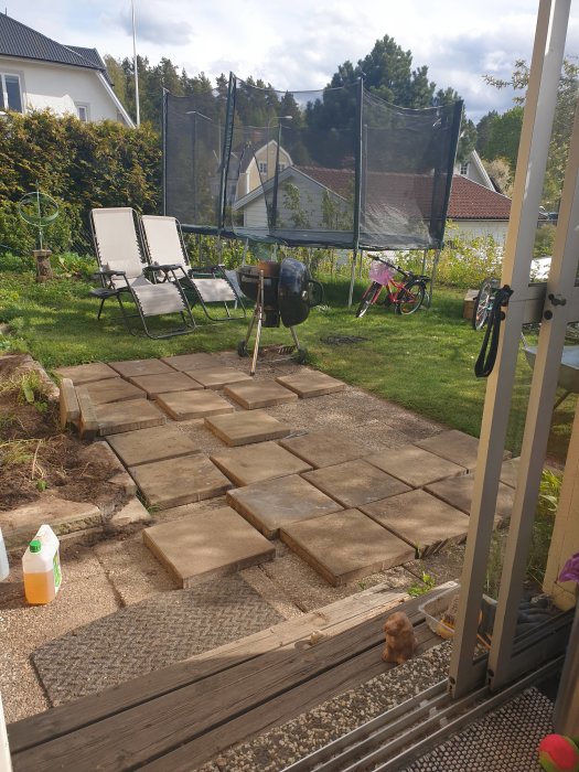 Nytvättade stenplattor ordnade i formation för en uteplats i en trädgård, med stolar och grill i bakgrunden.