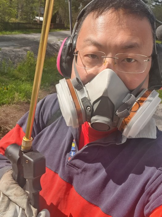 Person i arbetskläder med hörselskydd och andningsmask håller i en verktygsstång, förberedd för trädgårdsarbete.