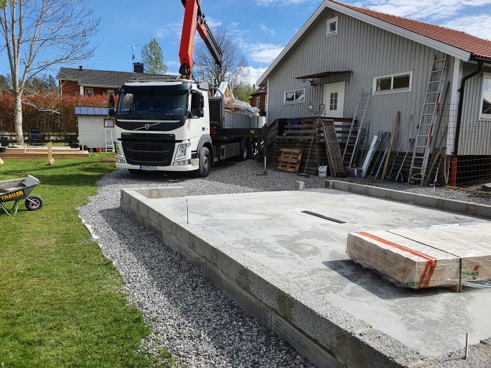 Lastbil levererar virke till en betonggrund för garagebygge med hus och trädgård i bakgrunden.