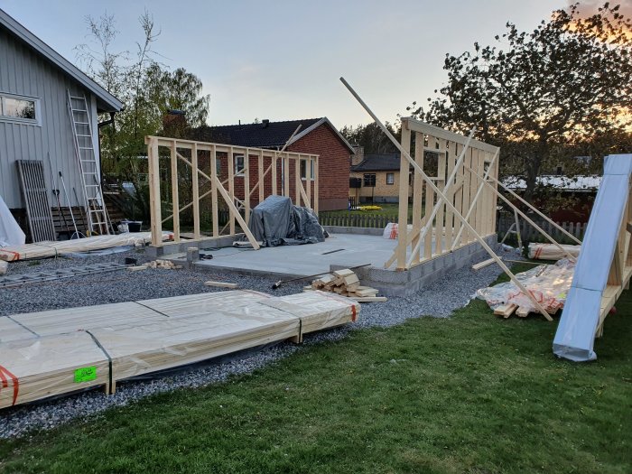 Byggstomme till ett garage under uppbyggnad med reglar och hammarband på en grundplatta, omgiven av oöppnat virke och verktyg.