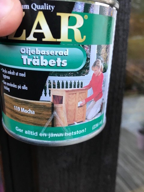 En hand som håller en burk med oljebaserad träbets, modell 119 Mocha, med en bild på hur det används.