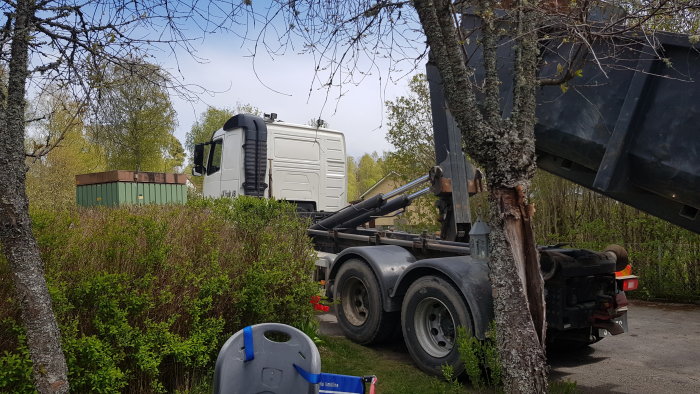 Lastbil med tippflak upp, boogieaxlar synliga, i en trädgårdsmiljö, antyder hög lastkapacitet.