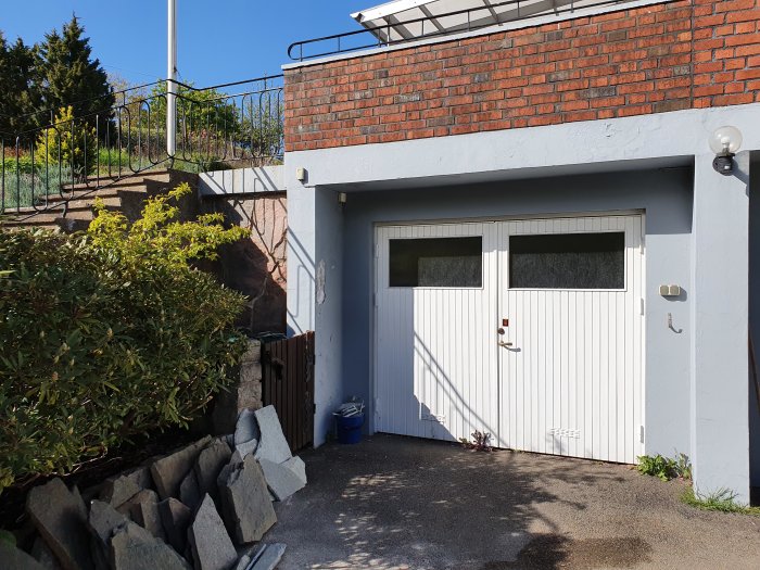 En husvägg med ett vitmålat garage under en tegelmur, där färgen ser ut att vara sliten och bubblig på grund av fukt.