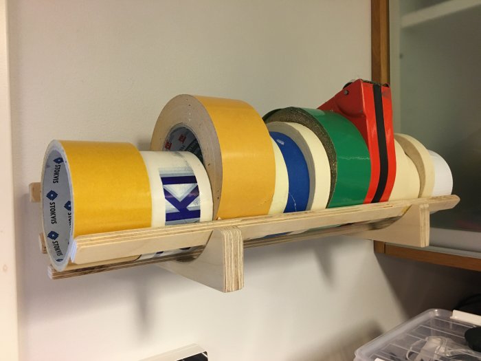 Hemmagjord hylla av plywood på vägg med olika rullar av tejp i varierande storlekar och färger.