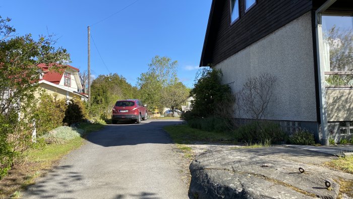 Utsikt över en uppfart med en röd bil, intillande hus och ytan där en pool och parkering är planerad.