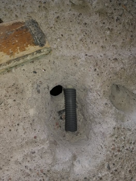 En flexslang installerad i ett betonghål, med synlig grusig yta och ett trästycke i övre vänstra hörnet.