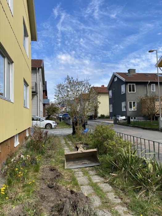 Gult tvåfamiljshus med grannhus och smidesstaket längs en smal uppfart och blommande trädgård.