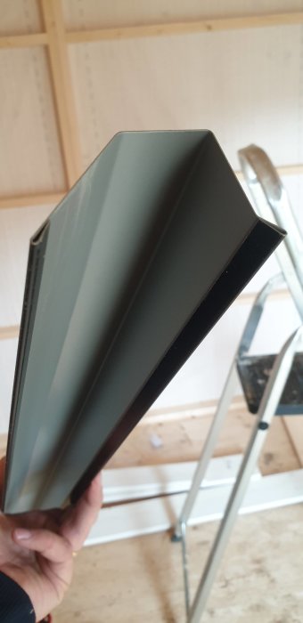 En hand håller upp en nockplåt för tak inuti en friggebod under konstruktion.