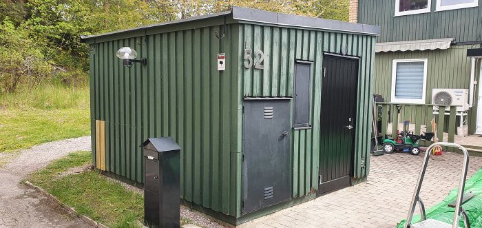 Grönmålad förrådsbyggnad med gammal panel från 1973, nyare ljusgröna panelbitar synliga, nära ett bostadshus.