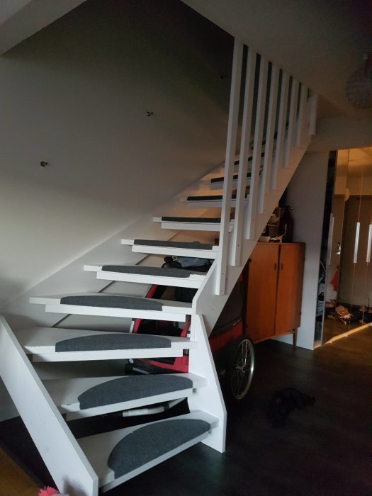 Vit trappa med svart spaljé som svänger 90 grader i ett hemmiljö med gråa trappstegsmattor.