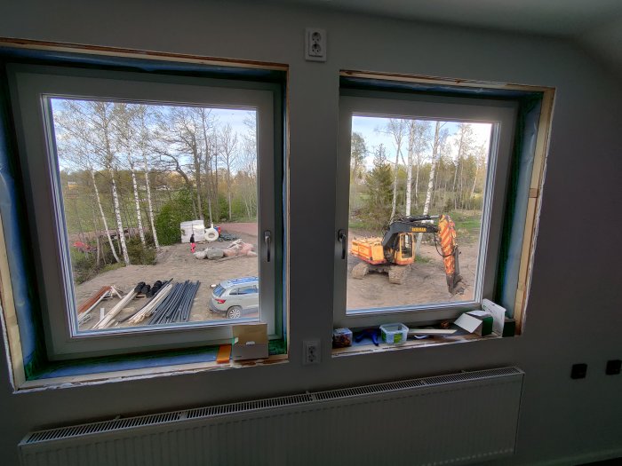 Utsikt från ett nybyggt hus med byggmaterial och en grävmaskin genom två fönster.