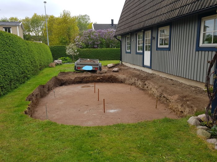 Grävt hål i trädgård med sandlager i botten och remsor runt förberett för poolinstallation.