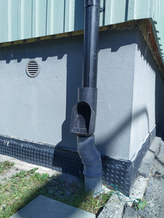 Stuprör kopplat till ett självrensande vattenutkast vid husgrund med synlig frostsprängning i putsen.