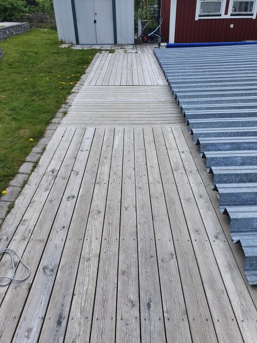 Slitet trädäck vid hus med gråa trallbrädor och delvis synlig trapp i metall.