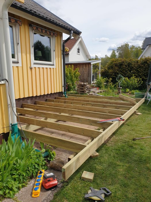 Konstruktion av en träaltan med reglar och plankor i en trädgård, byggarbete påbörjat bredvid ett gult hus.