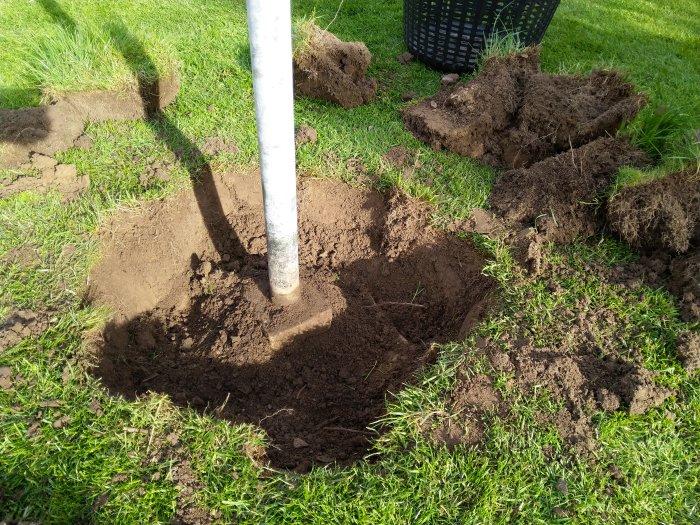 Uppgrävning av ett stålben förankrat i ett betongfundament i en gräsmatta, med jord och grästuvor runtomkring.