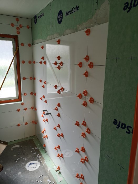 Badrum under renovering med vita kakelplattor och orange distansbrickor på väggen.