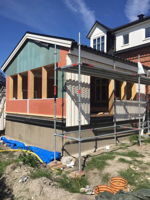 Obyggt hus tillbygge med reglar och spånplattor synliga, träfasad pågår, omgivet av byggställningar.