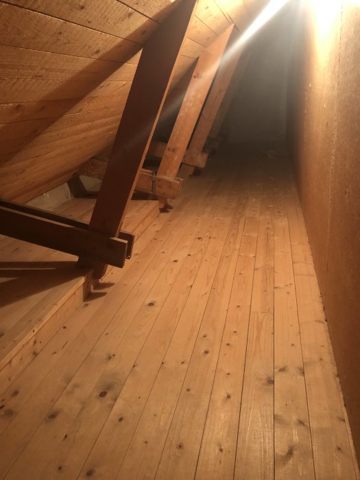 Träbjälkar och spånskivor på en kattvind med trägolv belyses av en ljusstråle.