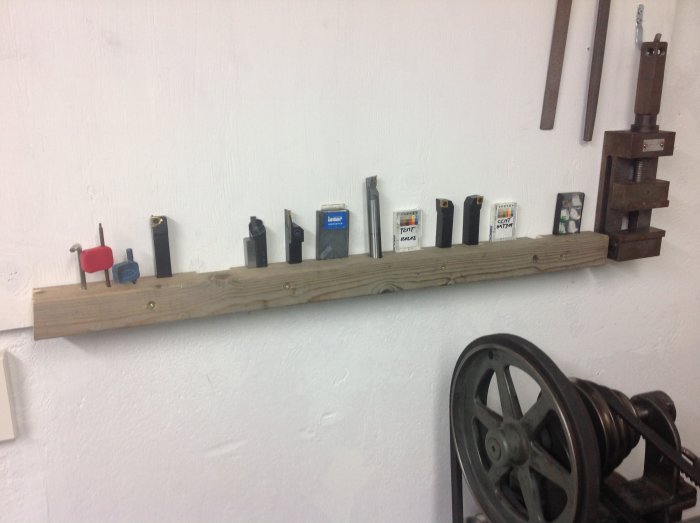 Hemmagjord verktygshållare med flera svarvverktyg och skärplattor ordnade på en trälist mot en vit vägg.