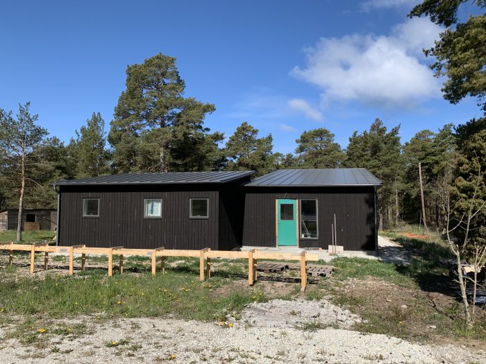 Nybyggt hus med mörkt tak och träfärgade väggar, grön helglasad ytterdörr, under en blå himmel.