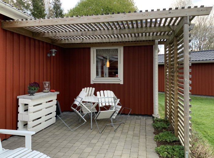 Röd stuga med pergola utan takmarkis, vitt trädgårdsmöblemang och gröna växter på uteplatsen.