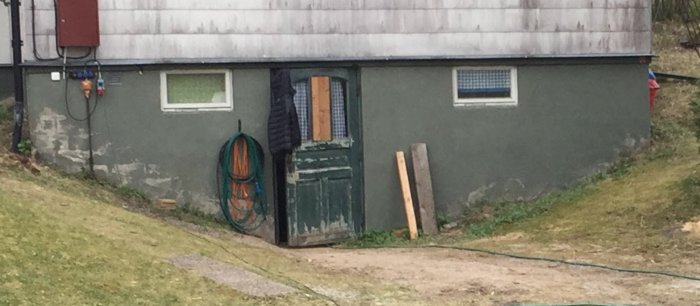 Utsidan av en källare med mörkgrönt mönster och eternittak, sladdar och en slangvinda synliga.