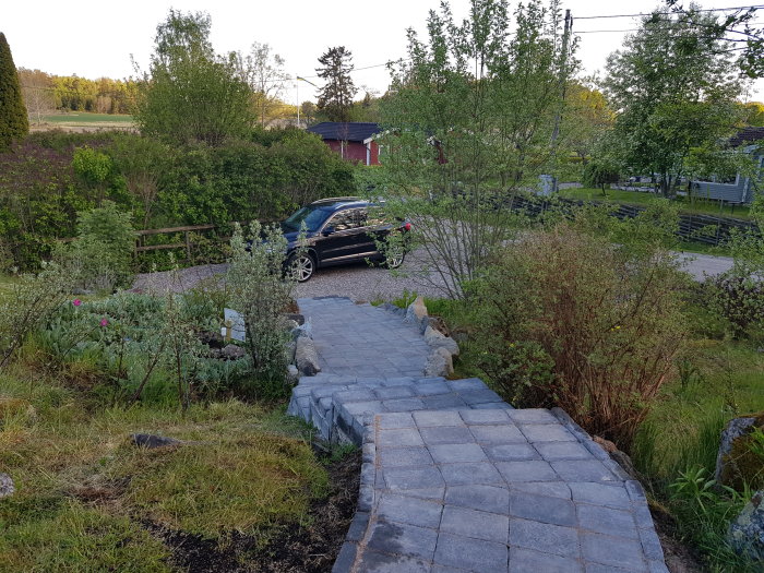 Stenlagd trädgårdsgång leder ner mot en bil parkerad vid ett staket, omgiven av växter och träd, med hus i bakgrunden.