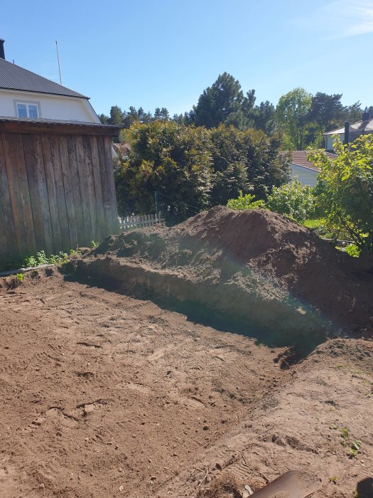 Stor jordhög bredvid utgrävningsgrop i trädgård med trästaket och hus i bakgrunden.