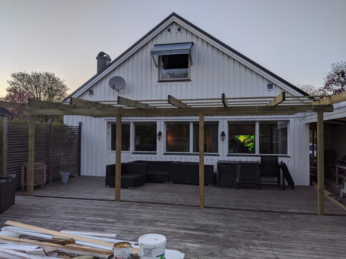 Altantak under konstruktion med stolpar och bärlinor på en trädäckterrass vid ett vitt hus.