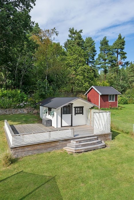 Fristående vit skjul bakom ett bostadshus med vedförvaring, omgiven av grönska.