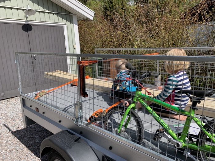 Två barn sitter i ett släp fäst vid cykel, säkrade med orange spännband, framför ett hus.