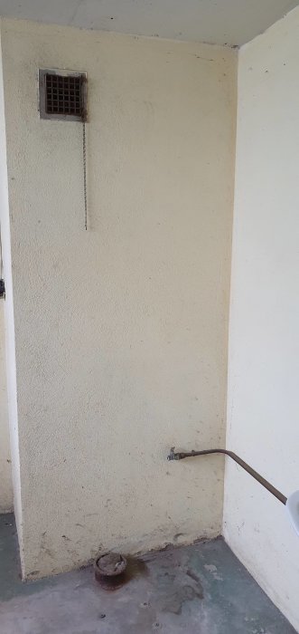 Hörn av en oinredd källare med en gammal ventilationsgaller högt upp på väggen och en avstängd vattenledning nära golvet.