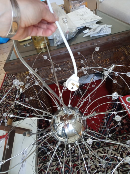 Ny taklampa med metallarmar och prydnadskristaller monterad ej, synlig elkontakt och installationsmaterial i bakgrunden.