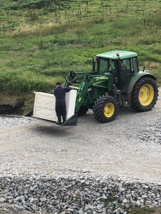 Person som använder en traktor för att lyfta en stor vit låda på ett grusunderlag.