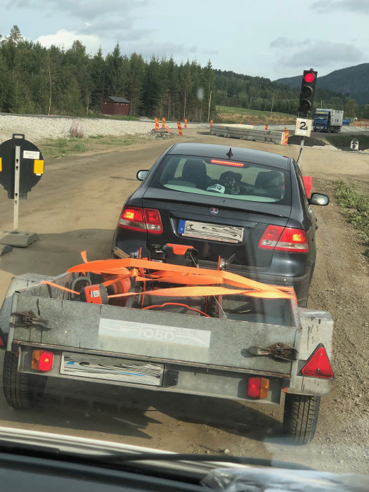 Bil med släp lastat med verktyg säkrade med orangea spännband, sett från ett fordon bakom vid vägarbete.