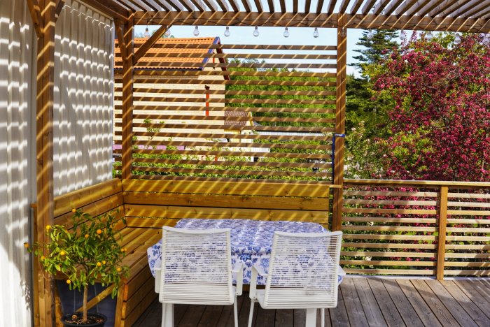 En trädäck med pergola och inbyggd soffa, ett blommande träd i kruka, och ett vitt bord med stolar.