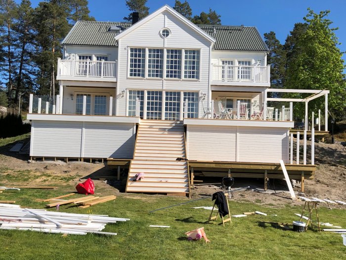 Vit villa med nya altaner på båda sidor och byggmaterial över gräsmattan, pergola under konstruktion åt höger.