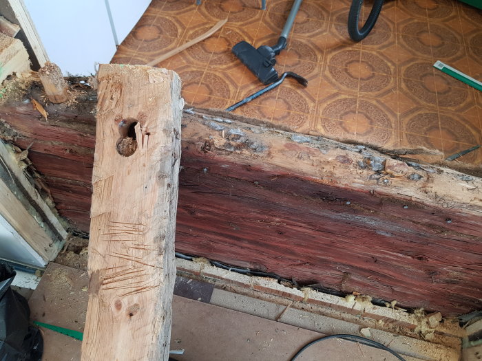 Skadad träbjälke med röta och en avsliten planka med hål, i ett pågående renoveringsprojekt.