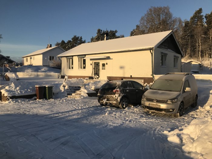 Vit husfasad och bilar täckta av snö på en solbelyst vinterdag.