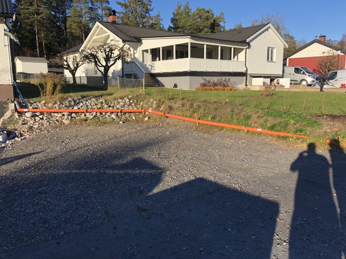 Uppgrävd trädgård vid vit husfasad med orange markränna under arbete, skuggor på grus.