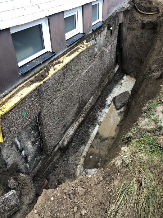 Grävd schakt vid husgrund med synlig felaktig dränering och vattenskador, samt betongrör och vattenansamling.