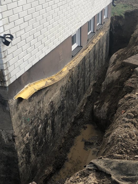 Grävningsarbete vid husgrund med synligt fuktskador, grävmaskinens grävning och vattenansamling i grop.