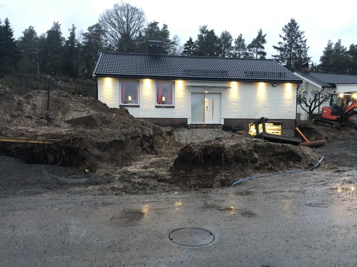 Hus med grävmaskin och uppgrävd mark runtom, synliga avloppsrör och provisoriska trappor till entrén.