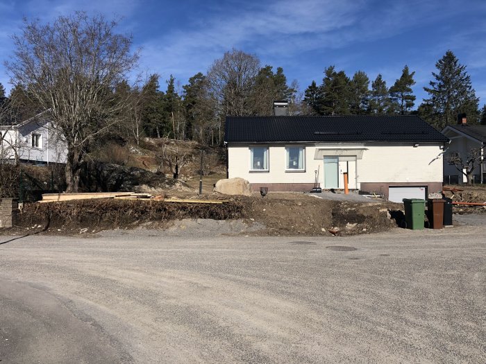 Renoveringsarbete på en ojämn tomt framför en vit enplansvilla med utspridda byggmaterial och verktyg under vårsolen.