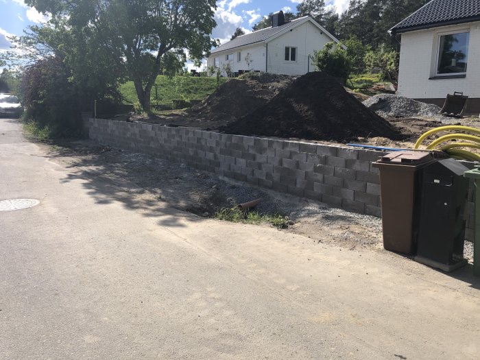 Nybyggd stödmur av gråa betongblock vid en tomt, med grushögar och anläggningsmaterial synligt.
