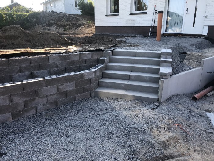 Stödmurar och trappsteg i betong under konstruktion vid ett hus, grus och byggmaterial synligt.