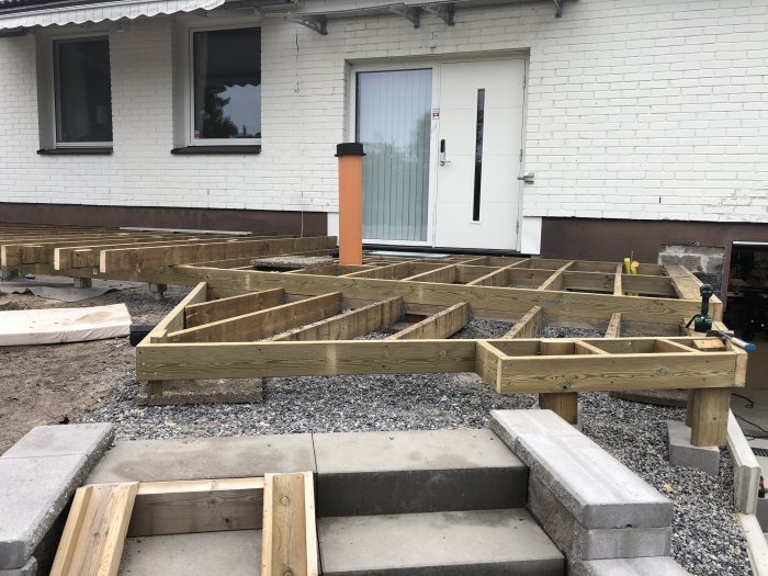 Grundstrukturen för en frukostaltan under uppbyggnad med distinkta vinklar utanför ett hus med vita tegelväggar.
